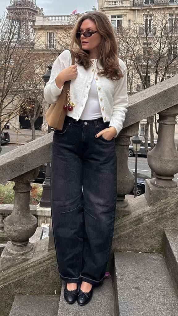 The outfit features a classic white cropped cardigan with delicate gold buttons paired with high-waisted, barrel leg jeans in dark denim, creating a modern yet timeless silhouette. This looks effortlessly chic and is complemented by sleek black flats, adding a touch of sophistication while maintaining comfort for fall and winter outings. Accessorized with a minimalist tan handbag, the ensemble balances casual elegance with a stylish edge, making it ideal for seasonal layering.