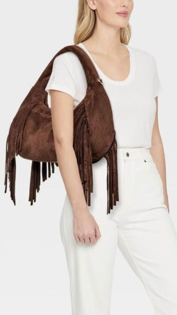 The outfit features a classic white crew-neck t-shirt paired with high-waisted white trousers, creating a fresh and clean silhouette. The standout element is a rich brown suede handbag adorned with long fringe, adding a touch of bohemian flair while remaining elegant and sophisticated. This combination balances modern trends with timeless style, perfect for the fall and winter seasons.