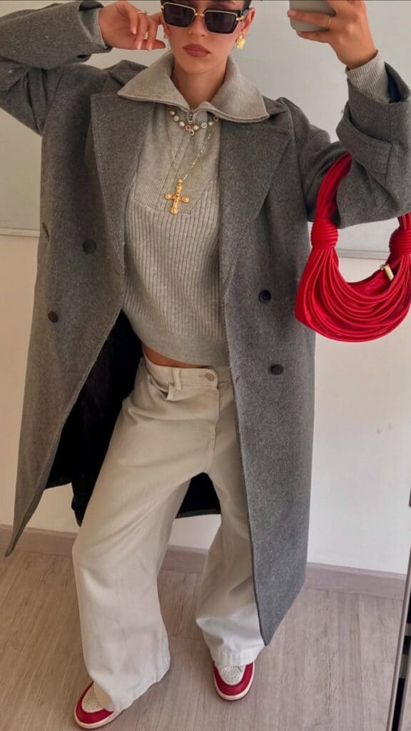 The outfit features a tailored gray overcoat layered over a soft, ribbed gray sweater, complemented by bone-colored wide-leg trousers that provide a relaxed silhouette. A pop of red is introduced through stylish sneakers with red accents and a bold red handbag, while gold jewelry adds a touch of elegance, creating a chic yet timeless look perfect for the fall and winter seasons. The overall vibe is effortlessly sophisticated, blending contemporary trends with classic elements.