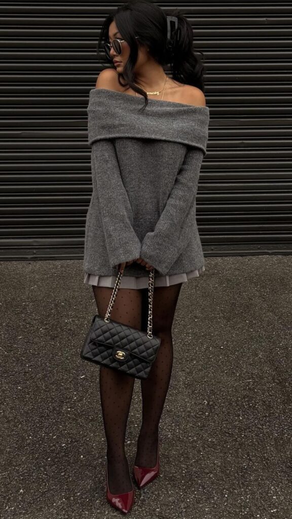 The outfit features an oversized, off-the-shoulder gray sweater that drapes elegantly, paired with a flared gray skirt that exposes a subtle layer of fabric. Sheer black tights adorned with delicate polka dots complement the look, while bold, glossy red heels elevate the ensemble further; a classic black quilted handbag adds a touch of sophistication. This combination creates a chic and timeless appearance, perfect for the fashion-forward individual navigating the fall and winter seasons.