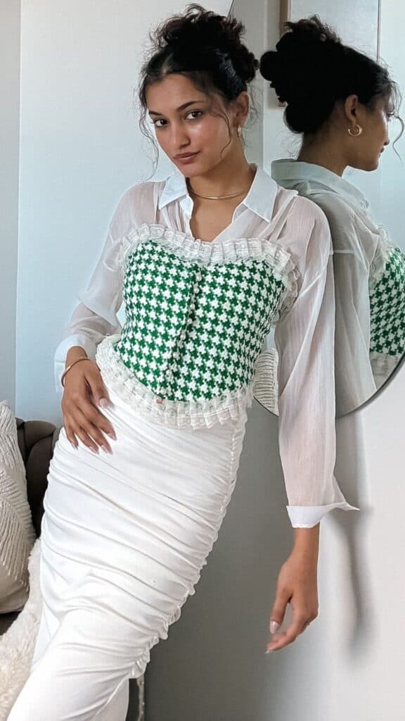 The outfit features a sheer, white button-up shirt layered under a modern Victorian-inspired corset, showcasing a vibrant green and white houndstooth pattern accented with delicate ruffles along the neckline. Paired with a form-fitting, ruched white skirt that elegantly drapes to highlight the silhouette, this ensemble effortlessly merges timeless elegance with contemporary style, perfect for the fall and winter seasons of 2025/2026. The overall vibe radiates sophistication, merging classic elements with a chic, modern twist.