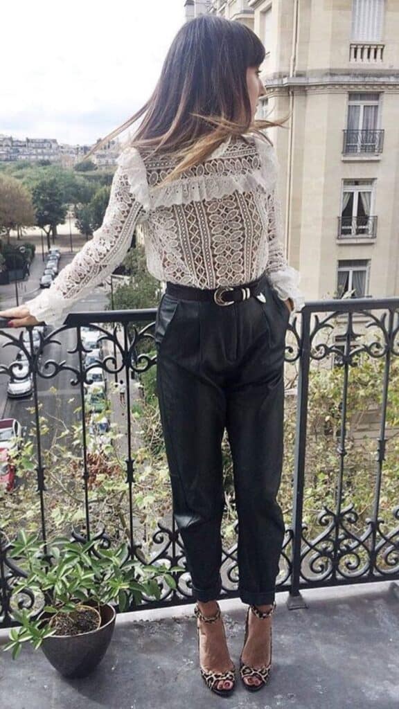 The outfit features a delicate, sheer lace blouse with intricate Victorian-inspired patterns and ruffled details at the cuffs and neckline, paired with high-waisted, tailored black leather pants that provide a modern contrast. This sophisticated ensemble is complemented by sleek, animal-print high-heeled sandals, creating a refined yet on-trend look that captures the essence of contemporary Victorian aesthetics while maintaining timeless elegance.