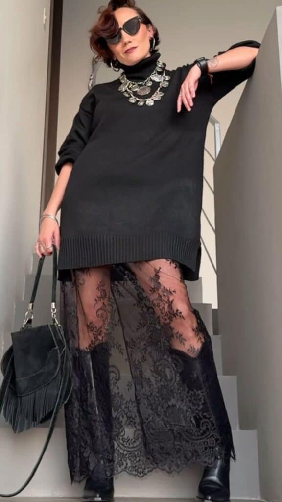 The outfit features a cozy black oversized sweater paired with a dramatic full-length lace skirt overlay, creating a feminine silhouette that blends casual and chic. The intricate lace detailing adds a touch of elegance, while bold accessories like a statement silver necklace and oversized sunglasses enhance the sophisticated vibe, perfect for fall and winter 2025/2026 fashion trends. This timeless look balances comfort and style, making it versatile for various occasions.