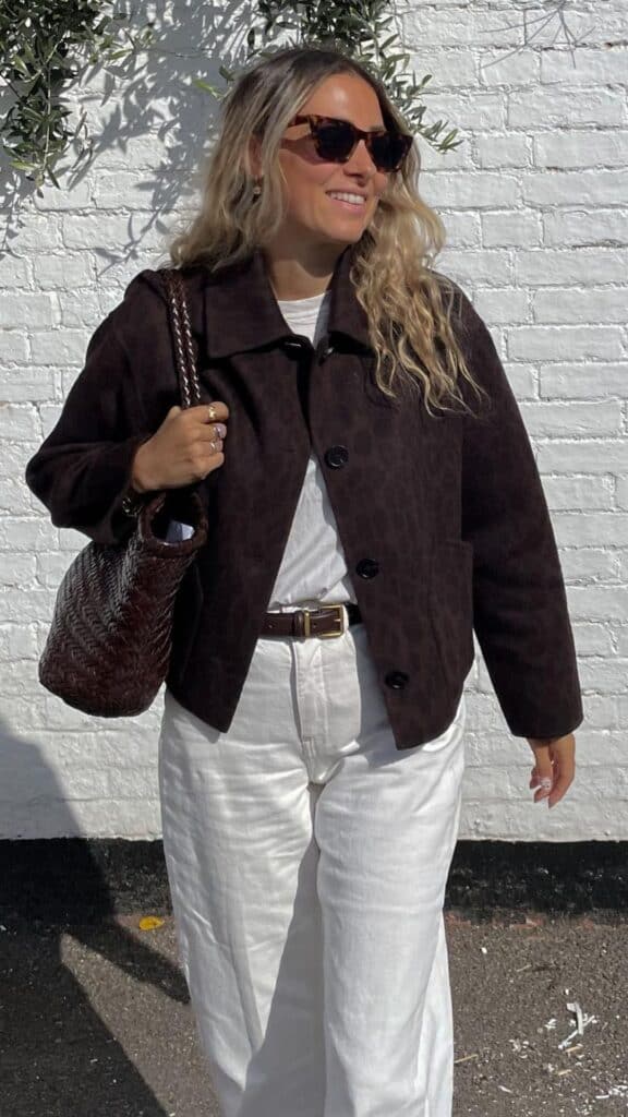 The outfit features a dark brown leopard print jacket, offering a subtle yet trendy animal print that adds depth to the look while maintaining a classic silhouette. Paired with high-waisted, off-white wide-leg trousers, the ensemble strikes a balance between bold and sophisticated, enhanced by a simple white top and accessorized with a woven, dark brown handbag. This combination creates a chic and timeless vibe, perfect for fall and winter 2025/2026.