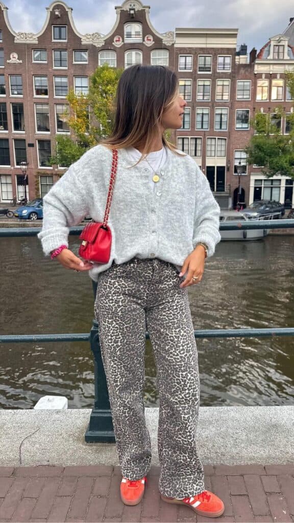 A cozy, light gray, oversized cardigan features a button-up design, layered over a classic white T-shirt, exuding effortless comfort and style. The eye-catching leopard print flared pants create a bold statement, while vibrant red sneakers and a matching small crossbody bag add a playful yet sophisticated touch overall, ensuring a chic and modern look for fall and winter.