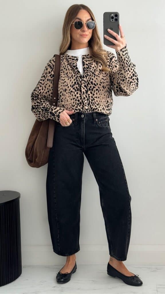 The outfit features a stylish leopard-print button-up shirt with voluminous sleeves, layered over a white top, paired with high-waisted barrel leg jeans in a deep black wash for a modern yet classic silhouette. Black flats with a delicate bow detail enhance the look, while a brown suede tote adds an element of warmth, creating a timeless and sophisticated vibe ideal for fall and winter trends.