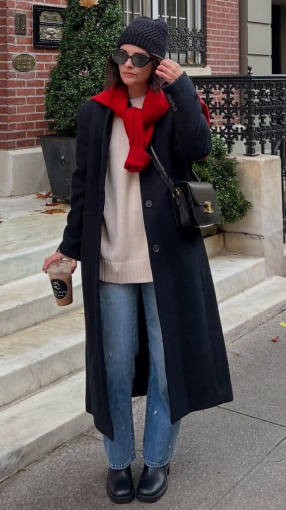 The outfit features a long navy coat over a cream ribbed sweater, complemented by relaxed-fit faded denim jeans, creating a sophisticated yet casual winter look. A bright red scarf adds a pop of color while enhancing the timeless feel, while black ankle boots and a sleek handbag complete the ensemble, ensuring it's both stylish and practical for the season's trends.