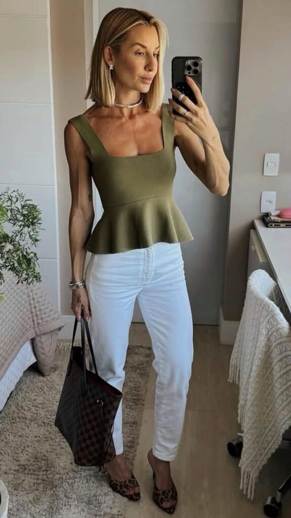 The outfit features a fitted olive green peplum top that accentuates the waist with a structured silhouette, paired with sleek white high-waisted jeans that create a sharp contrast. The look is completed with stylish leopard print heels and a chic, dark tote bag, exuding an elegant yet contemporary vibe perfect for fall and winter. The ensemble balances modern trends with timeless pieces, making it suitable for various occasions.