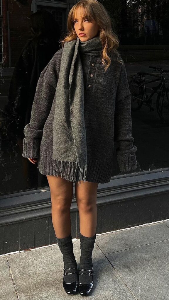 An oversized charcoal knit turtleneck sweater features a relaxed fit with dropped shoulders and a buttoned neckline, exuding a cozy, yet chic vibe for the fall and winter season. Paired with a long, fringed gray scarf that adds texture, the outfit is accessorized with knee-high ribbed socks and sleek black patent leather Mary Jane shoes, creating a balance between comfort and style that feels both timeless and elegant. This ensemble embodies the trend of oversized knitwear while maintaining a sophisticated aesthetic, perfect for seasonal wear.
