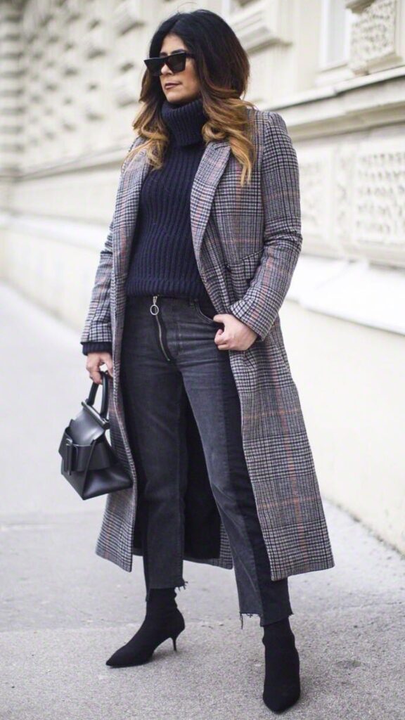 The outfit features a tailored, long plaid overcoat in soft grays and hints of orange, layered over a cozy, navy turtleneck sweater, creating a sophisticated contrast. The ensemble is completed with high-waisted, dark gray straight-leg jeans that highlight ankle-length black sock boots, emphasizing the current revival of this trendy footwear. A structured black handbag adds a modern touch, resulting in a chic, timeless look perfect for the transitional fall and winter seasons.