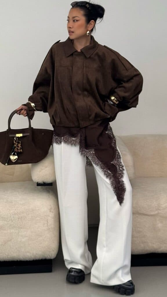 The outfit features an oversized, rich brown suede jacket with a structured collar and relaxed fit, paired with high-waisted, flowing white trousers that elongate the legs. A delicate lace detail in a darker brown cascades from the jacket, adding a feminine touch, while chunky black platform shoes provide a modern, grounded finish, creating a chic and timeless look perfect for fall and winter.