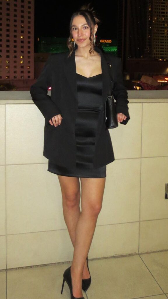 The outfit features a sleek, black satin mini dress that hugs the body and has a flattering, low neckline. Complementing this, a long, tailored black blazer adds sophistication while providing warmth, and the entire look is elevated with pointed black stiletto heels. Accessories include a small black handbag, contributing to a polished and elegant appearance suitable for outings like birthday parties or date nights.