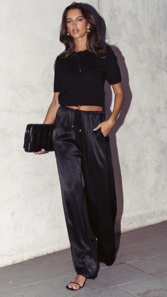 The outfit features a fitted black cropped cardigan with short sleeves, made from a soft knit fabric that complements the body. Paired with high-waisted, flowing black satin pants, the ensemble exudes elegance and sophistication, perfect for evening occasions. Accessorized with a structured black clutch and minimalist strappy heels, this look is ideal for birthday parties or date nights.