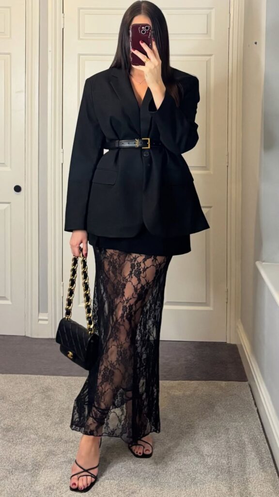 This elegant black outfit features a tailored blazer made from structured fabric, cinched at the waist with a black belt, creating a flattering silhouette. Underneath, a floor-length black lace skirt reveals a glimpse of skin, enhancing the feminine touch, and the look is completed with strappy black high-heel sandals and a small chain handle handbag. The combination of textures and the layered design adds sophistication, making it perfect for evening events and special occasions.