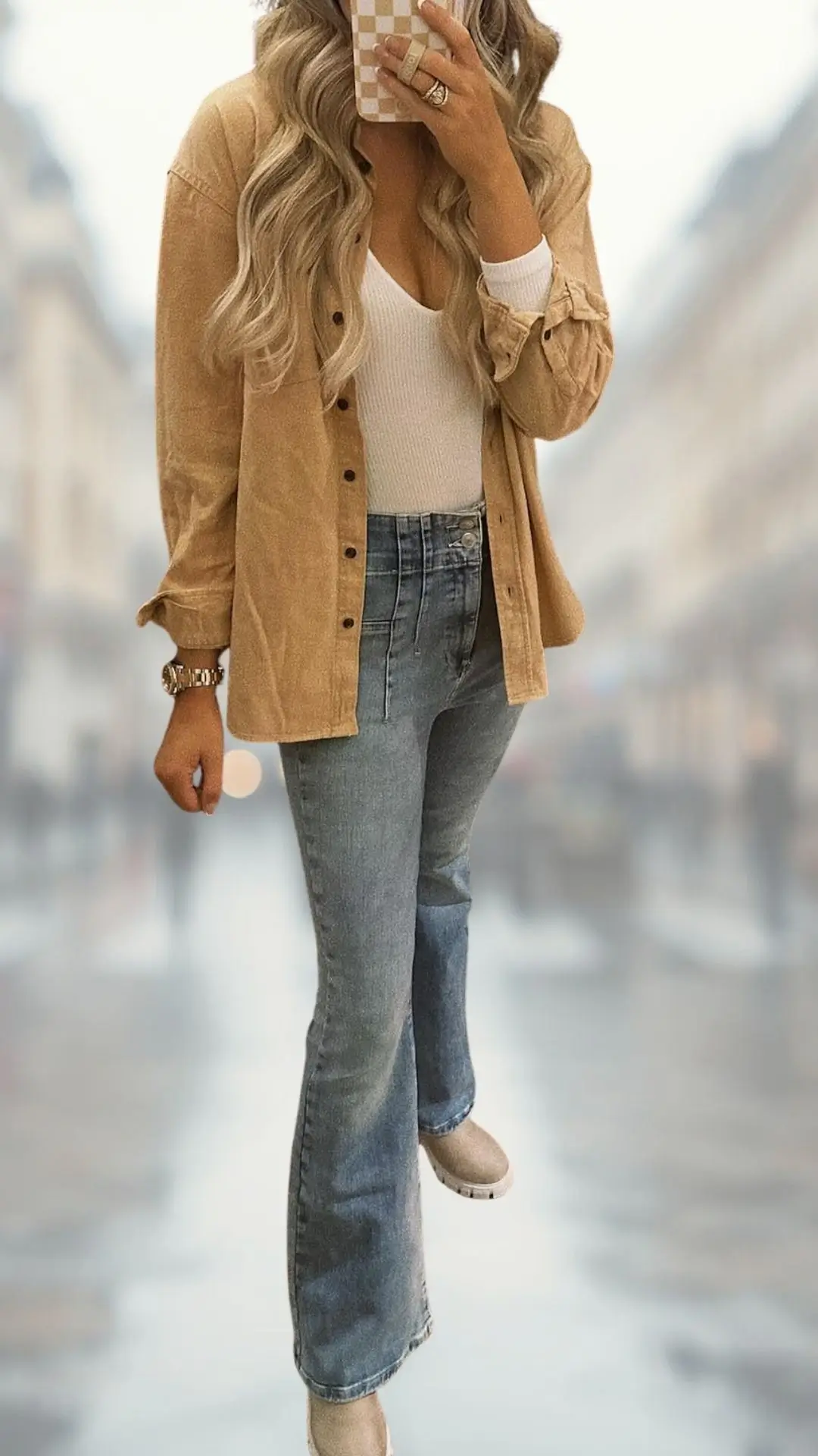 The outfit features a light beige corduroy button-up shirt layered over a fitted white ribbed tank top, paired with high-waisted blue flare jeans that accentuate the legs. The ensemble is completed with stylish, light-colored ankle boots that add a touch of sophistication. The combination of textures and soft shades creates a balanced and fashionable look.