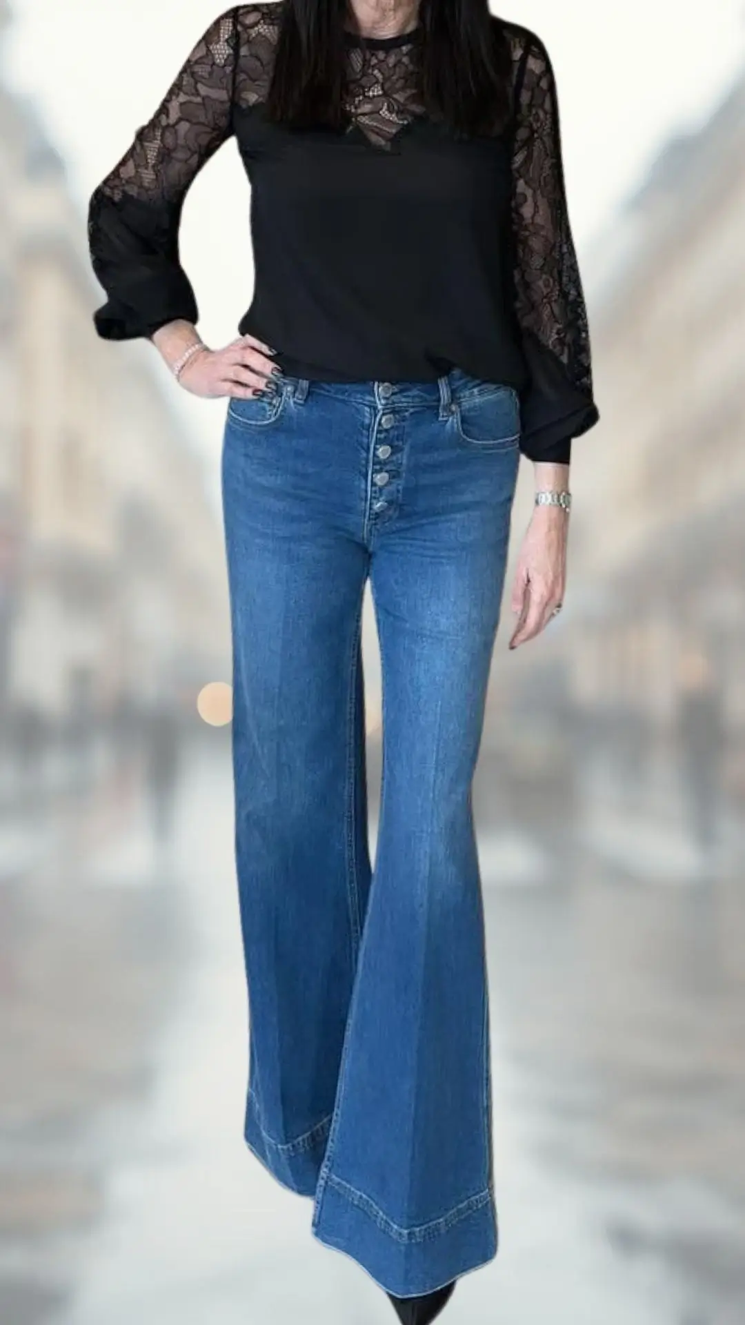 A stylish outfit features a black blouse with sheer lace long sleeves and a rounded neckline, paired with high-waisted, flared denim jeans in a medium blue wash. The jeans showcase a fitted silhouette at the top and gradually widen at the hem, while the look is completed with sleek, black ankle boots for an elegant finish.