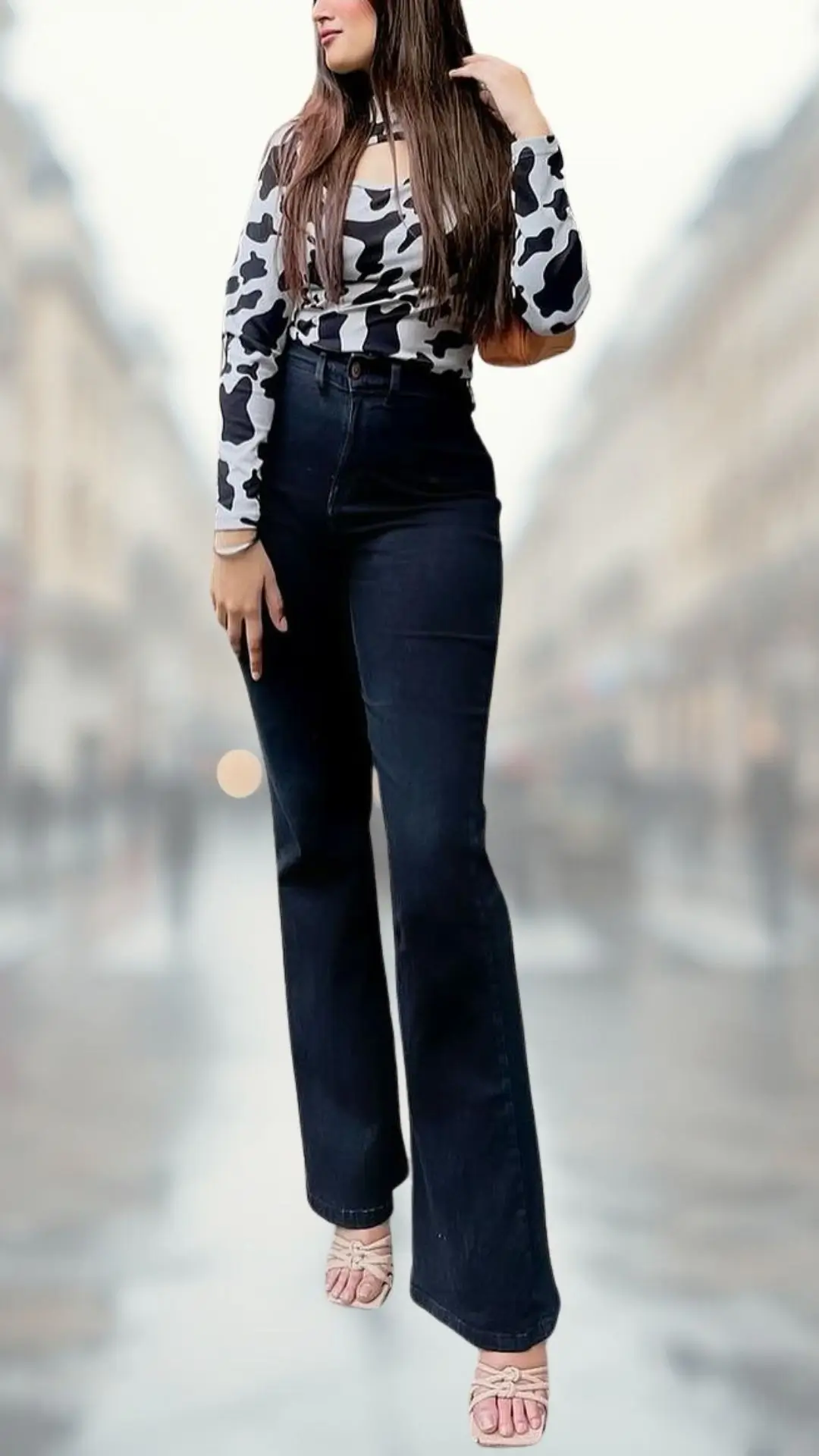 The outfit features a fitted long-sleeve top in a bold black and white cow print with a scoop neckline and cut-out detail, paired with high-waisted dark denim flare jeans that enhance the silhouette. Completing the look are strappy nude block-heeled sandals that add elegance and a touch of height. The overall combination of textures and shades creates a stylish, contemporary look suitable for various occasions.