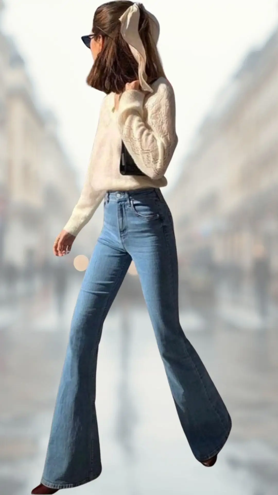 A woman is wearing a cozy, cream-colored knit sweater with long sleeves and a relaxed neckline, paired with high-waisted flare jeans in a light blue wash that elongate the legs. She completes the outfit with brown pointed-toe boots and accessorizes with a black clutch and a beige hair scarf tied in her hair. The overall look is stylish and classic, blending textures and shades harmoniously.