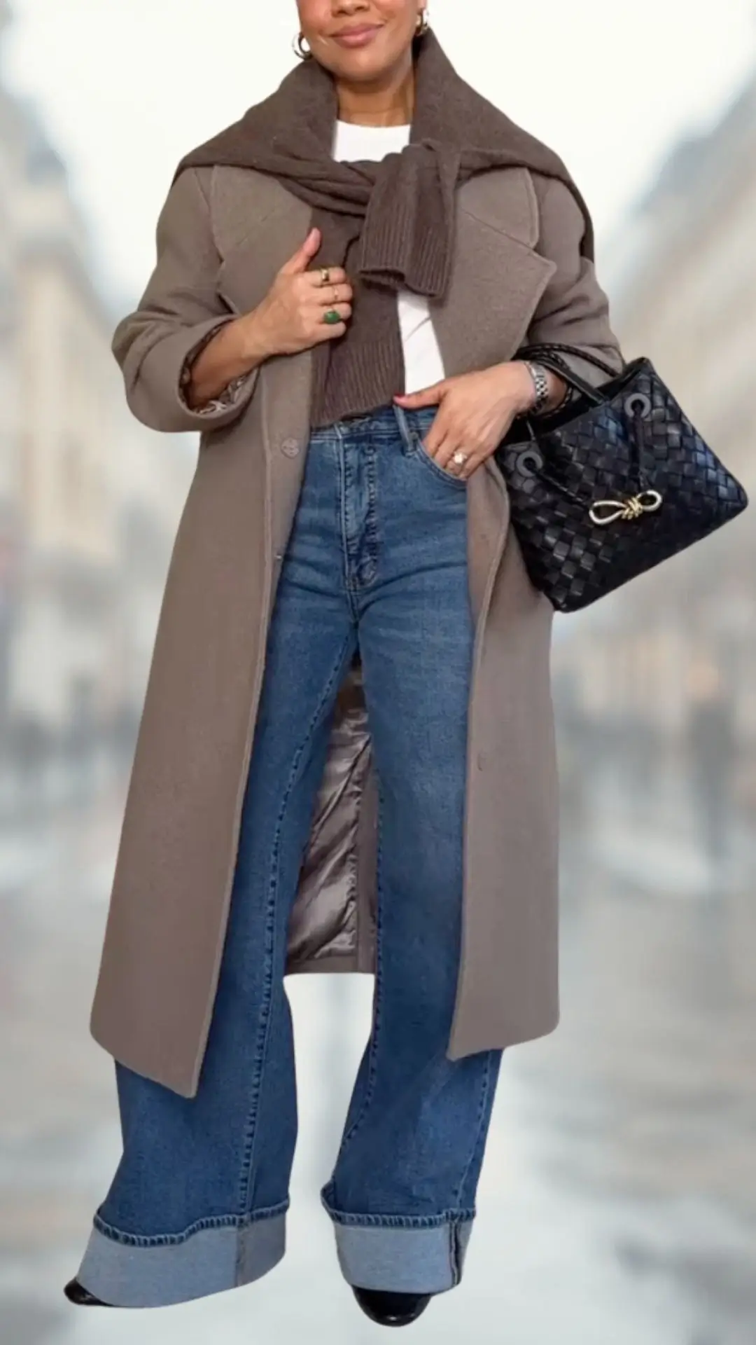 The outfit features high-waisted flare jeans in a rich blue denim, complemented by a cream-colored fitted top layered with a cozy brown sweater draped around the shoulders. A stylish taupe overcoat adds elegance, while black, pointed-toe heels and a quilted black handbag complete the look. The combination of textures and shades creates a refined and chic appearance.