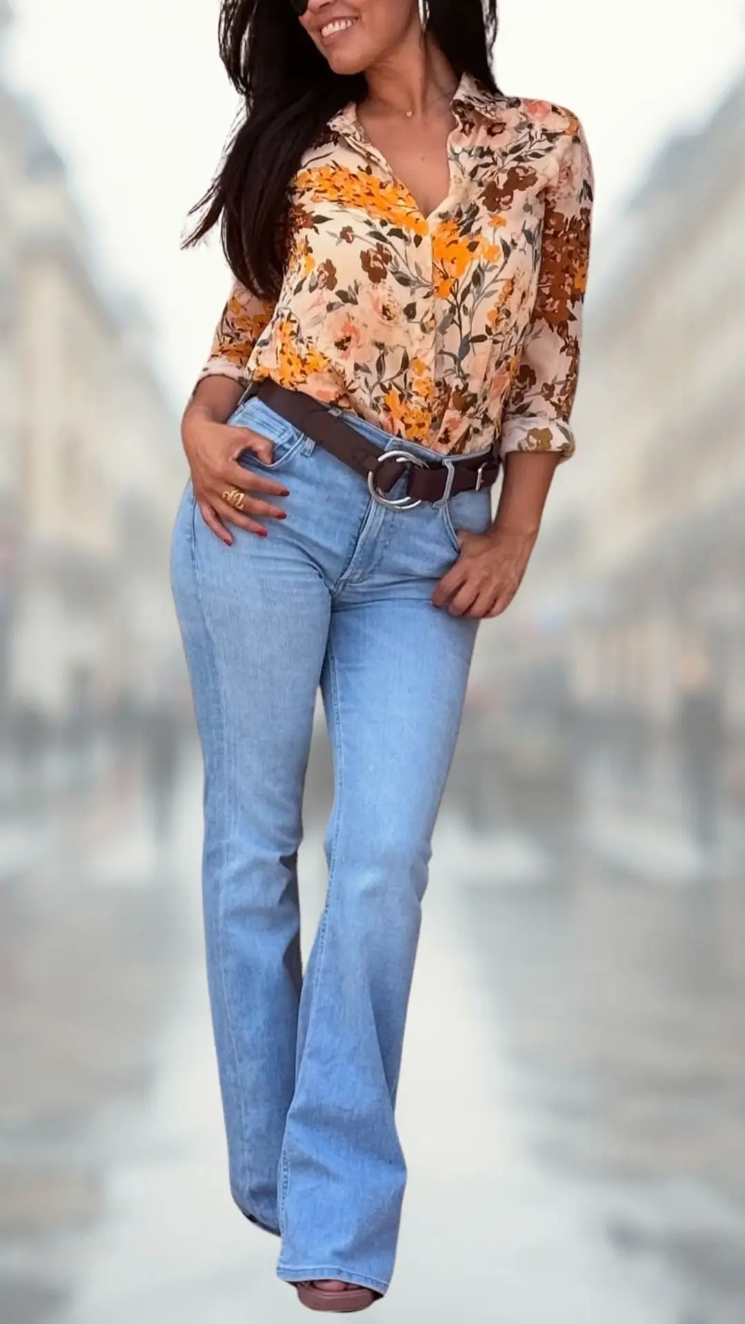 The outfit features a floral button-up shirt in warm shades of orange and cream, with three-quarter sleeves and a relaxed fit. Paired with light blue flare jeans, the look is completed with a brown leather belt and stylish sandals, blending comfort and elegance effortlessly. The combination of textures and colors creates a vibrant and cohesive aesthetic.