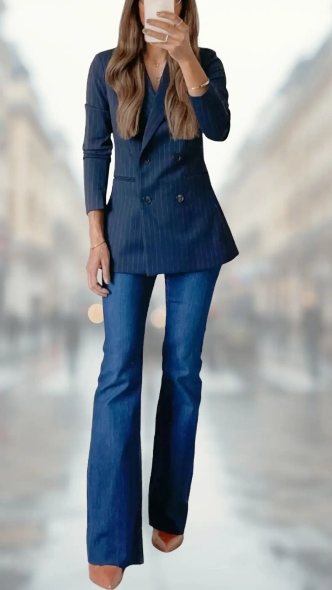 The outfit features a tailored, navy pinstriped blazer with long sleeves and a deep V-neckline, paired with flared dark denim jeans that gently distort into a soft bell shape. The look is completed with elegant nude high-heeled shoes that add height and sophistication. The combination of structured and casual elements creates a chic balance suitable for various occasions.