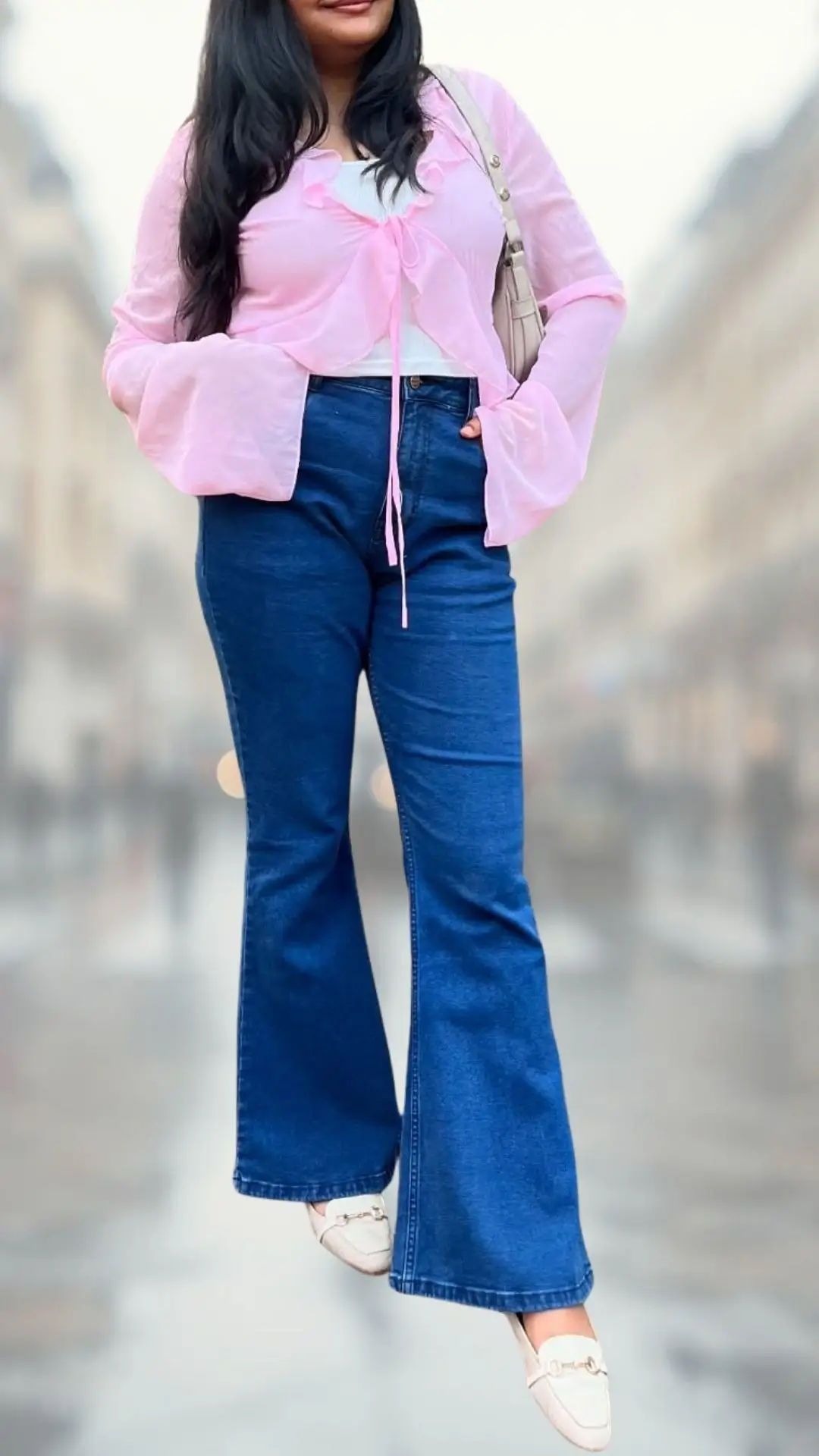 A flowy pink sheer top features bell sleeves and ties at the neckline, layered over a white fitted tank, paired with high-waisted dark blue flare jeans that extend to the floor. The outfit is completed with light-colored loafers adorned with a decorative buckle, creating a stylish and comfortable look for any occasion.