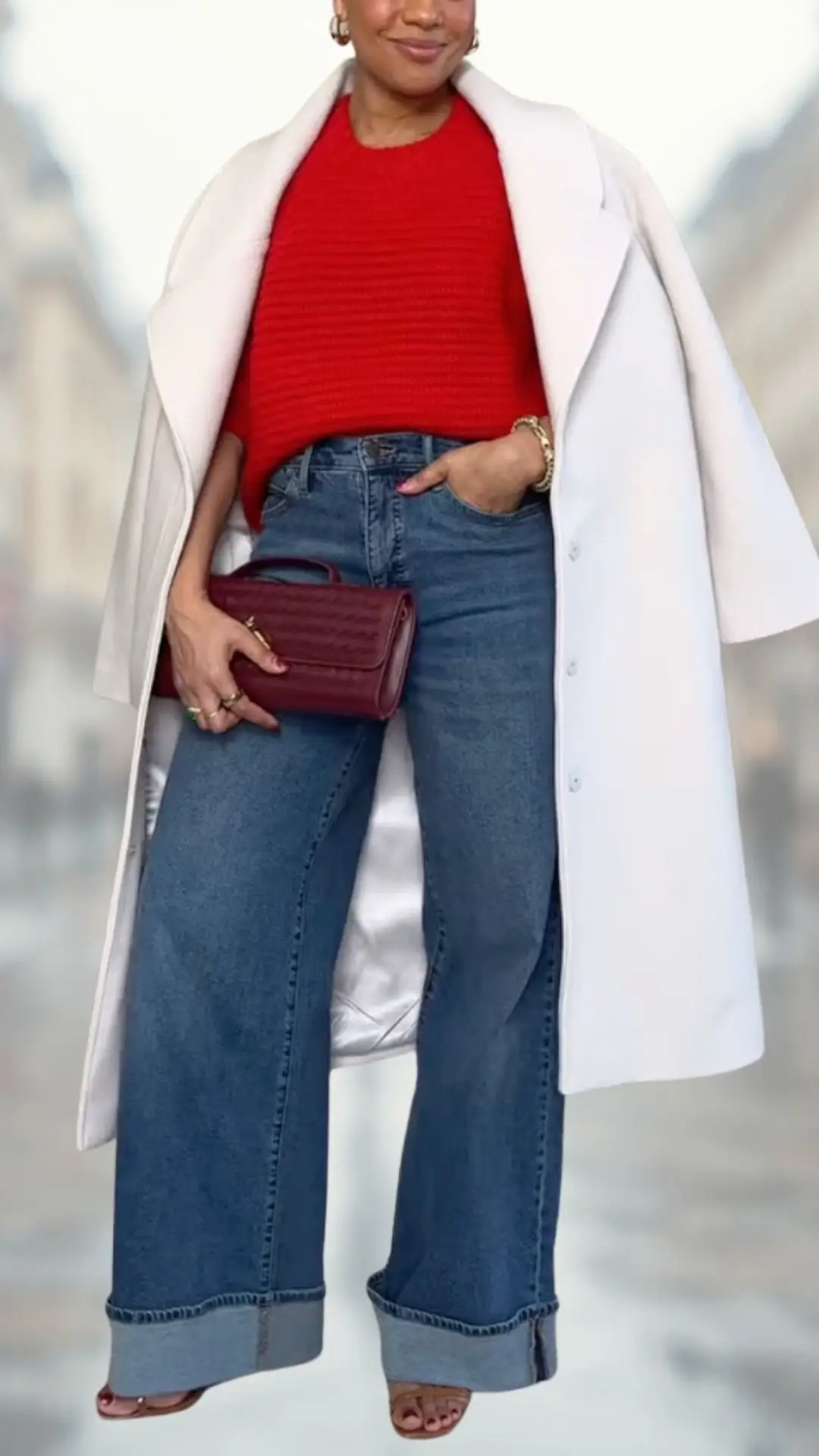 A vibrant red knit sweater features a round neckline and short sleeves, paired with high-waisted blue flare jeans that have a soft, denim texture and rolled cuffs. The outfit is completed with a chic white overcoat and accessorized with a small burgundy handbag and stylish open-toe heels, emphasizing a blend of comfort and elegance.