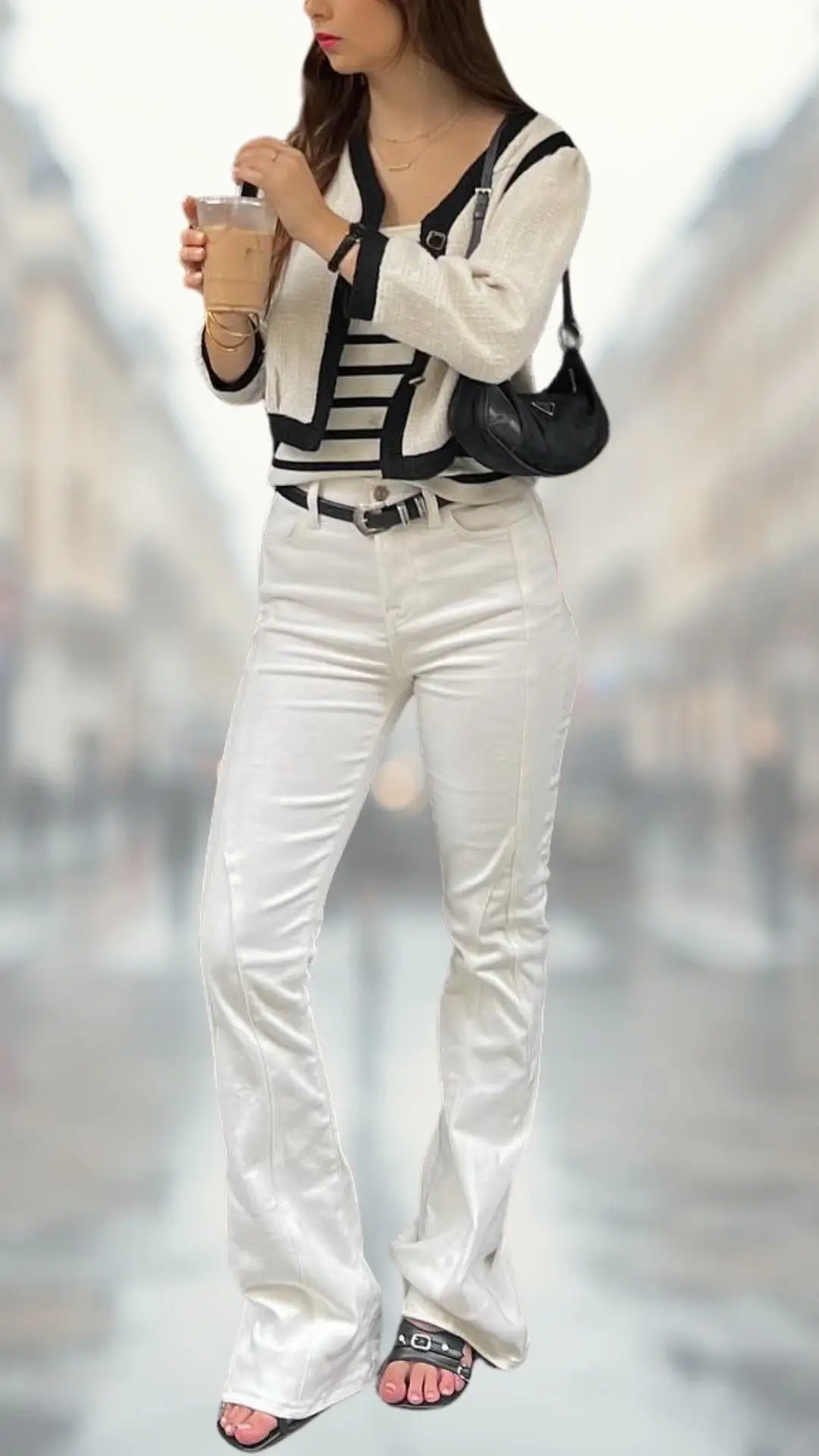 The outfit features a fitted black-and-white striped long-sleeve top, complemented by elegant white flare jeans crafted from a sleek fabric for a polished look. The ensemble is paired with stylish black slide sandals that have a buckle detail, and the model holds a drink, adding a casual yet chic touch to the overall aesthetic. A small black handbag completes the outfit, enhancing its contemporary vibe.