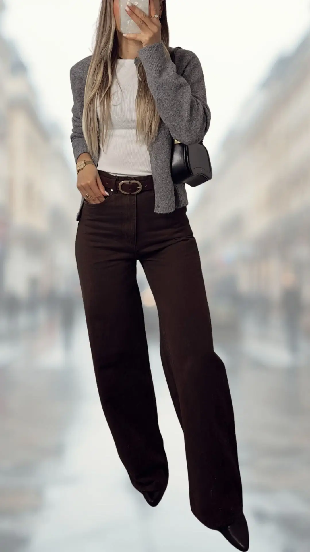 The outfit features a fitted white ribbed tank top paired with a soft grey cardigan, complemented by high-waisted brown flare jeans made from a sturdy fabric, creating a chic silhouette. An elegant black belt cinches the waist, and pointed black heels add a polished touch, enhancing the ensemble's sophistication. The look is both stylish and comfortable, perfect for a casual day out.