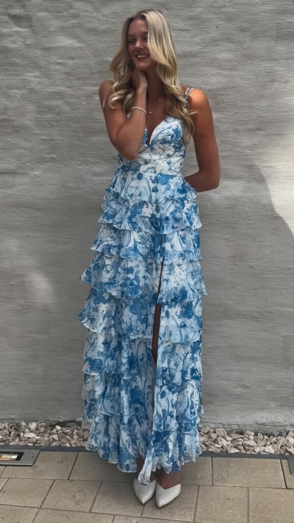 The dress features a vibrant blue floral pattern depicting delicate blooms and flowing vines, creating a soft, romantic aesthetic. It is a long, tiered gown with ruffled layers that enhance its femininity, complemented by a plunging neckline and a thigh-high slit for an elegant, flattering silhouette. This design is perfectly suited for wedding guest attire, exuding charm and grace.