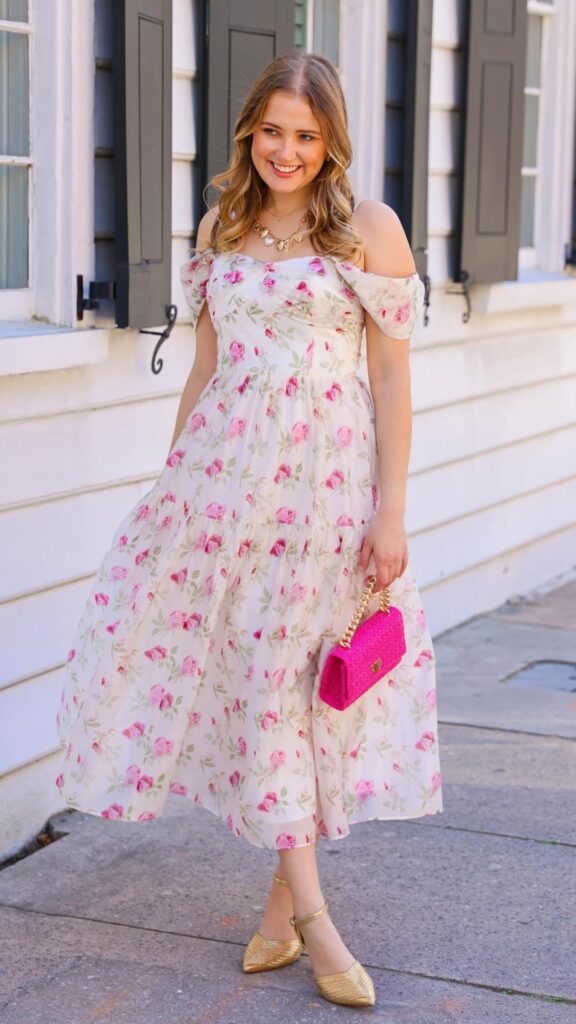The dress features a delicate floral pattern with soft pink roses and green leaves against a light, neutral background, evoking a romantic aesthetic. Its off-the-shoulder design and flowing maxi length create a flattering, feminine silhouette, complemented by a ruffled bodice and a tiered skirt that enhances movement. The look is elegantly accessorized with a bright pink clutch and gold heeled shoes, making it perfectly suited for a wedding guest.