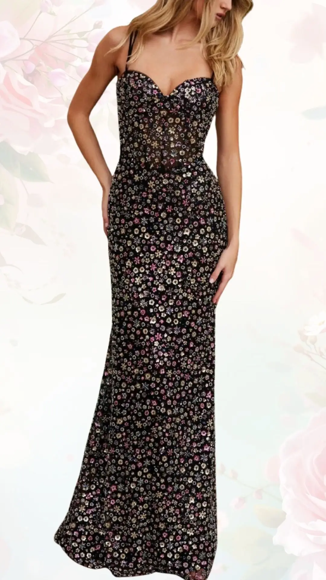 A fitted floral wedding guest dress features a sweetheart neckline and thin straps. The dress is adorned with a vibrant arrangement of small, colorful flowers against a dark black fabric, creating a striking contrast. It flows elegantly down to the floor, enhancing its formal appeal.