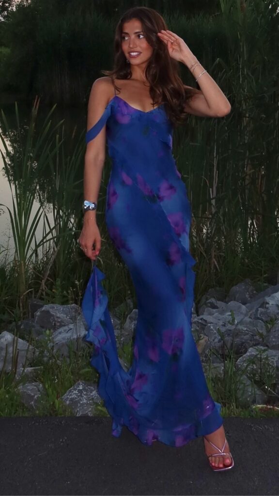 The vibrant blue floral wedding guest dress features soft, watercolor-like blooms in rich purples, elegantly cascading down the flowy fabric. With its refined spaghetti straps and delicate ruffles along the hemline, the dress beautifully enhances a feminine silhouette, making it a perfect choice for a festive and romantic occasion. Its lightweight material allows for graceful movement, contributing to an overall sense of elegance and charm.