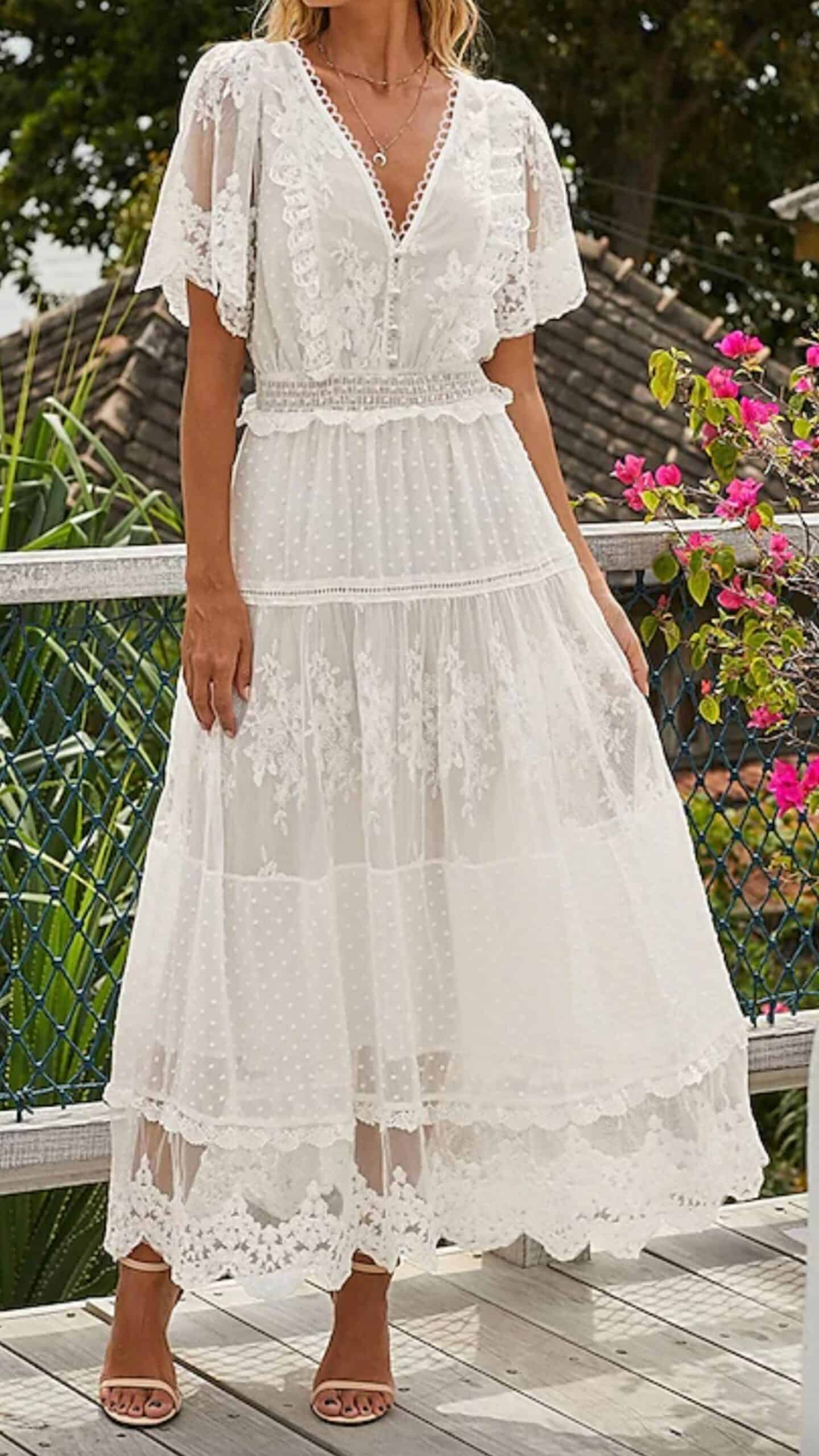 A flowy, full-length white dress featuring a deep V-neckline and flutter sleeves, crafted from lightweight, sheer fabric with subtle polka dots and delicate floral lace embellishments. The design includes a smocked waist and intricate lace trim at the hem, exuding a romantic vibe perfect for a summer wedding or garden party.