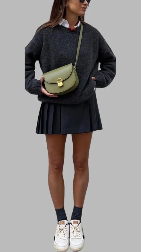 The outfit features a dark grey oversized sweater with a round neckline and long sleeves, paired with a black pleated skirt that falls above the mid-thigh. Complementing the look, the individual wears white sneakers with black accents and dark grey ankle socks, accessorized with an olive green crossbody bag. The overall ensemble balances comfort and style, showcasing a modern, casual aesthetic.