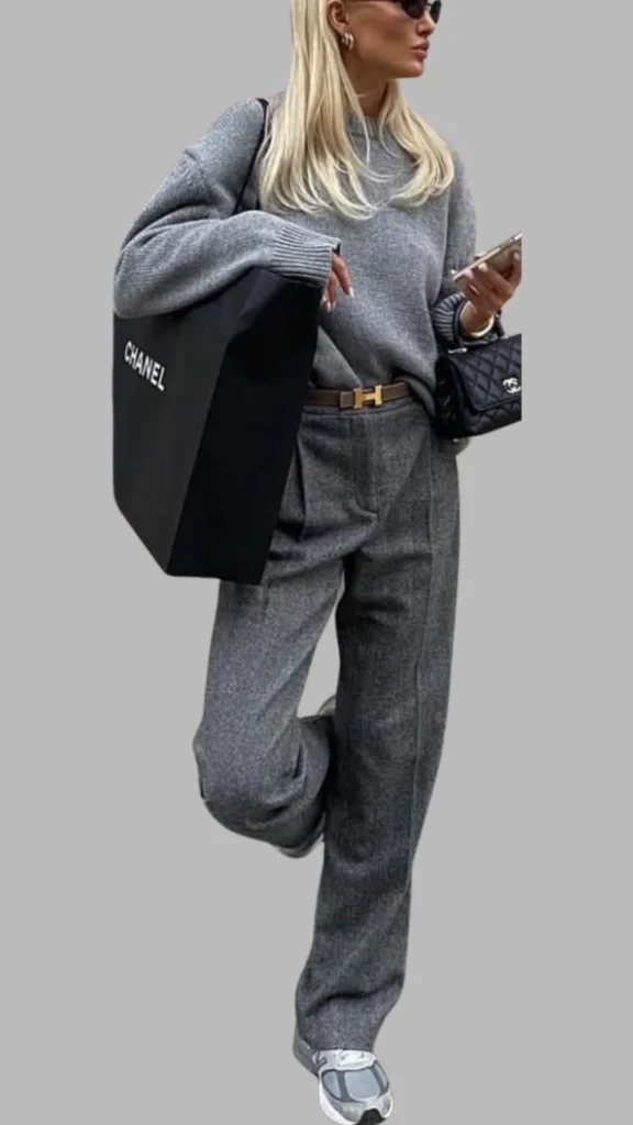The outfit features a relaxed-fit, light grey sweater paired with high-waisted, loose-fitting grey trousers, both made from textured fabric. The look is accentuated with a brown belt and completed with chunky grey sneakers, while a large black bag adds a chic contrast. The overall palette consists of various shades of grey, creating a sophisticated yet casual ensemble.