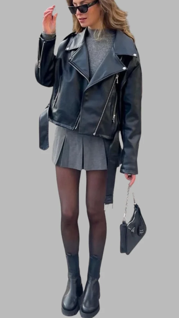 The outfit features a layered look with a fitted grey sweater peeking out from under a cropped black leather biker jacket, which has an asymmetric zipper and wide lapels. The dress underneath is a pleated, short grey skirt that adds a dynamic feel, complemented by sheer black tights and chunky black ankle boots for a stylish edge. The overall palette is monochromatic, with varying shades of grey and black, creating a sleek and modern aesthetic.