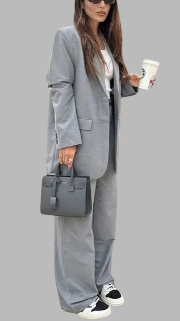 The outfit features a tailored grey oversized blazer paired with matching wide-leg trousers, creating a chic and modern silhouette. Underneath, a simple white tee adds contrast and balance, while the look is accessorized with a sleek black handbag and casual black and white sneakers, enhancing its versatility. The overall ensemble combines neutral tones with a polished yet relaxed vibe, perfect for various occasions.