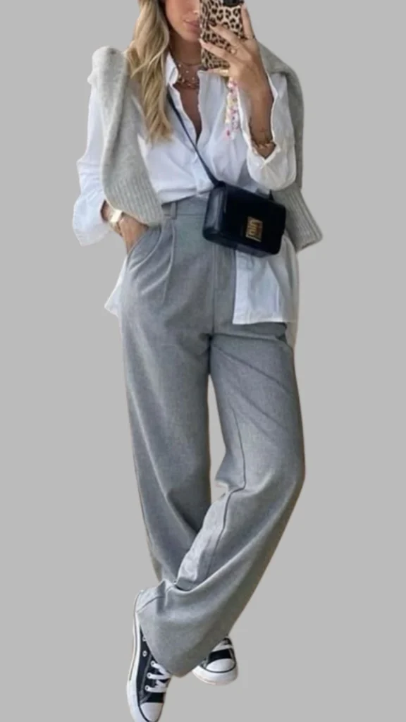 The outfit features wide-legged, light grey trousers with a high waist and a relaxed fit, paired with an off-white collared shirt that peeks out at the bottom. Layered on top is a soft, light grey knitted vest, creating a sophisticated yet casual look. The ensemble is completed with classic black and white sneakers for a modern touch.