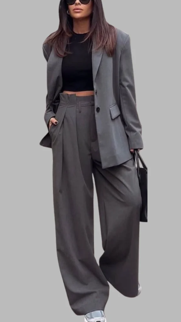 The outfit features a tailored grey blazer paired with high-waisted, wide-leg trousers, creating a sophisticated and stylish silhouette. The blazer has a notched lapel and a single button closure, while the trousers include subtle pleats and pockets for added functionality. This ensemble is complemented by a fitted black crop top, resulting in a chic, monochromatic look that balances elegance with modernity.
