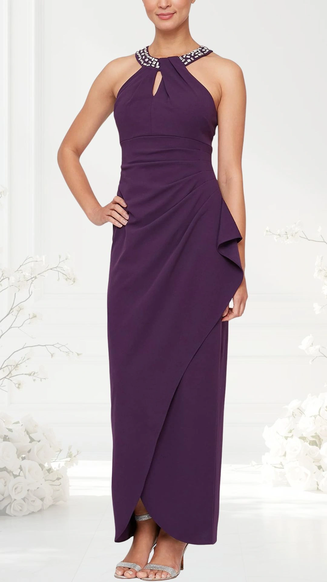 A halter neck wedding guest dress in rich purple features a shimmering beaded neckline and a keyhole detail at the front. The dress is elegantly form-fitting, with a draped side that creates a flowy silhouette, complemented by strappy silver heels. The background is a soft, light-colored space adorned with white floral accents.