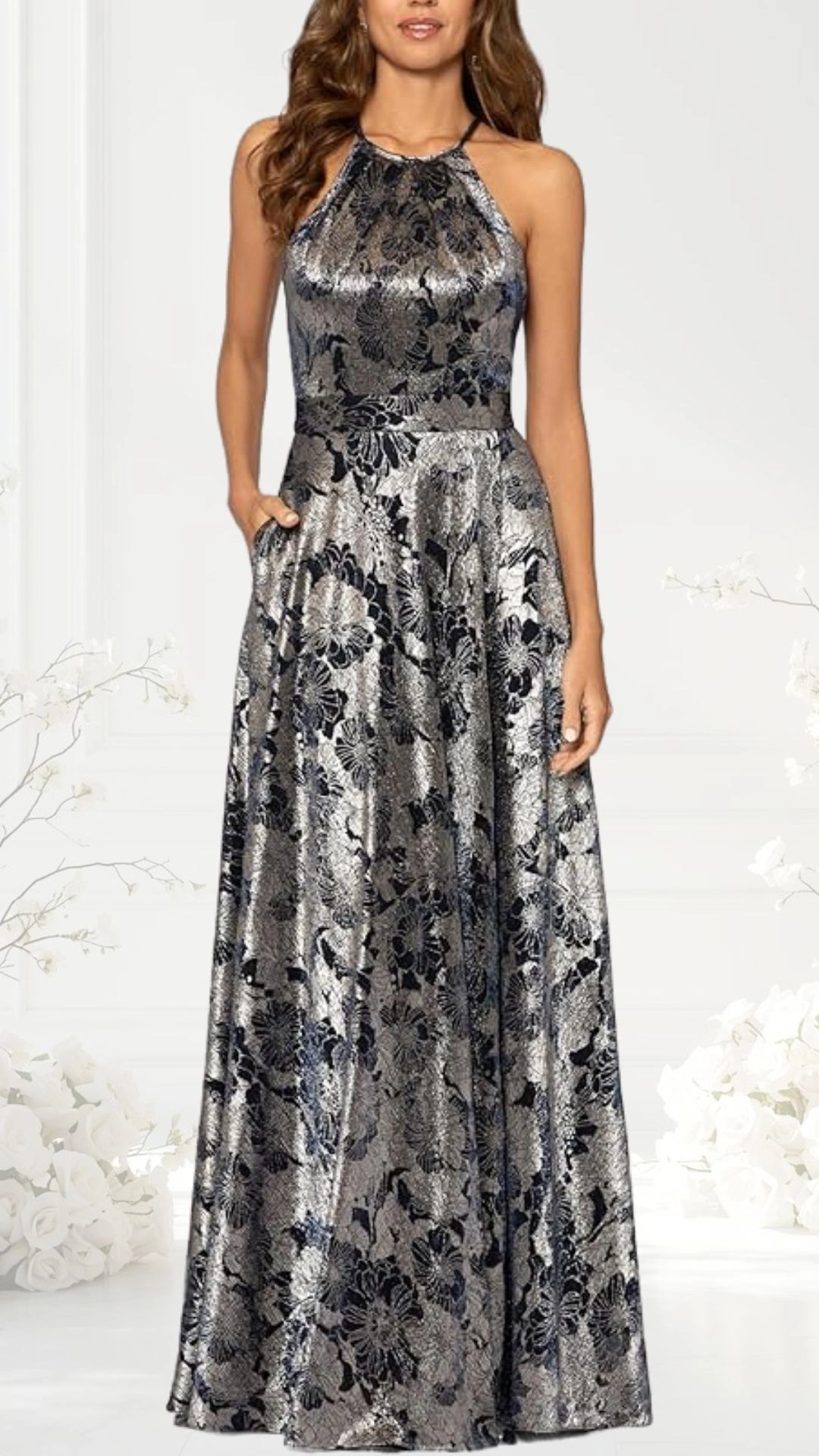 A halter neck wedding guest dress features a fitted bodice and a flowing, floor-length skirt, adorned with a shimmering floral pattern in shades of silver and navy. It includes pockets on either side for added convenience, creating an elegant silhouette. The dress is set against a soft, neutral background, enhancing its visual appeal.
