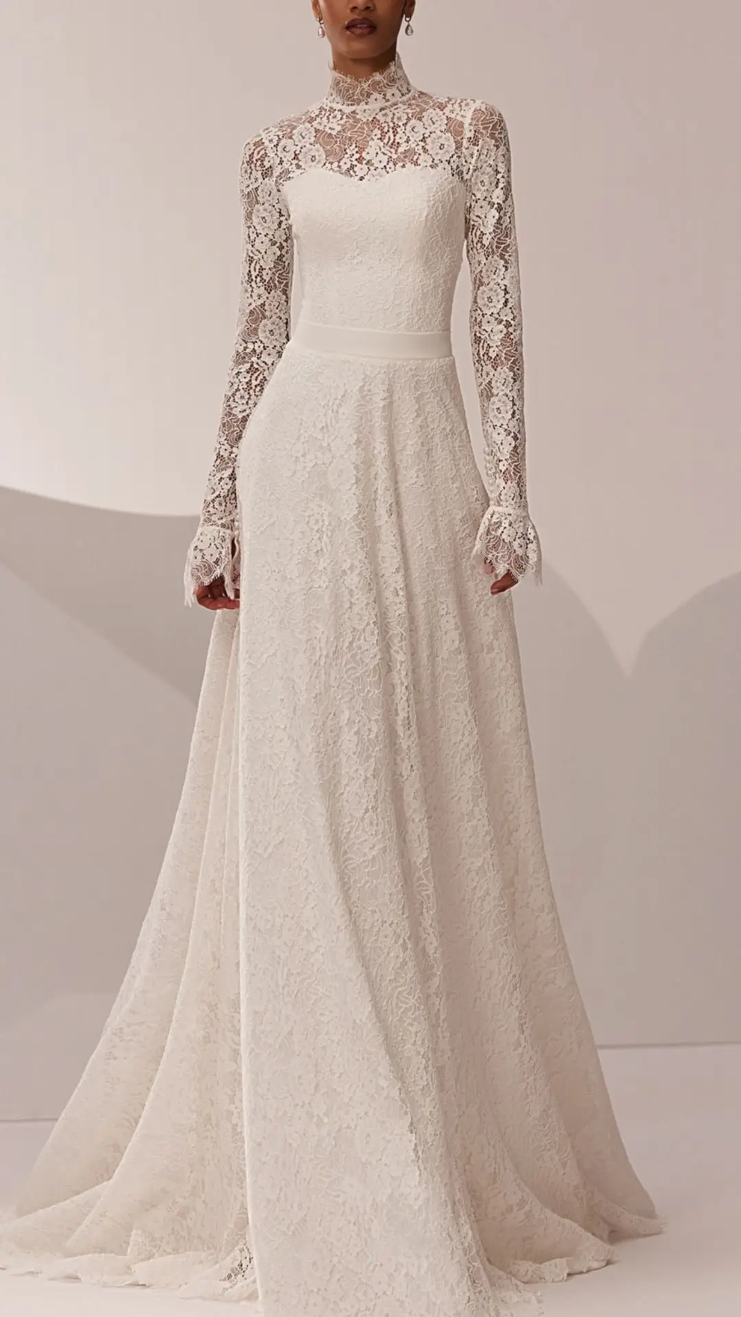 This high neck wedding gown features an elegant lace overlay with long, fitted sleeves that culminate in delicate lace cuffs. The dress is crafted from a soft, ivory fabric, creating a modest silhouette that flows into a graceful skirt, perfect for a winter wedding or vintage-inspired ceremony. The combination of the ornate lace and classic lines offers a timeless, sophisticated look.