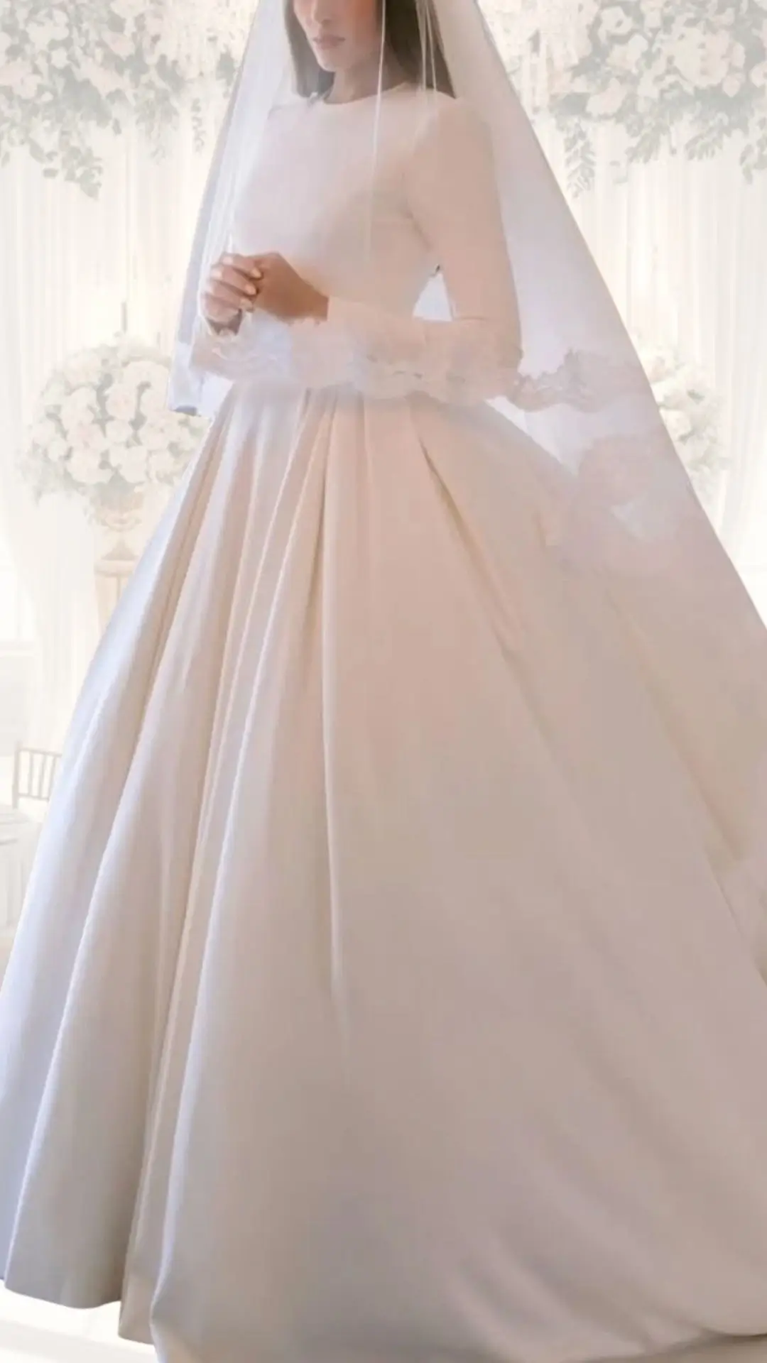 A high neck wedding gown featuring long sleeves and a flowing A-line skirt, crafted from luxurious off-white satin for a timeless elegance. The sleeves are accented with delicate lace cuffs, while the full skirt is elegantly pleated, making it ideal for a winter wedding or vintage-inspired look. A soft sheer veil complements the outfit, adding a classic touch.