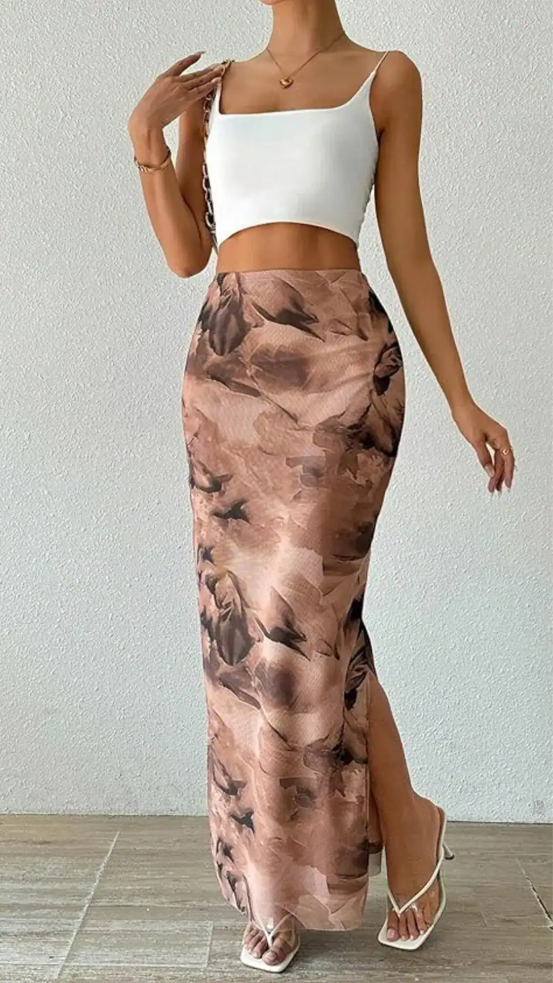 The outfit features a high-waisted floral midi skirt made from a lightweight, silky fabric, showcasing a dusty rose background adorned with dark, abstract floral patterns. Paired with a fitted white cropped top with a square neckline, the overall vibe is effortlessly chic and suitable for casual outings or summer events.