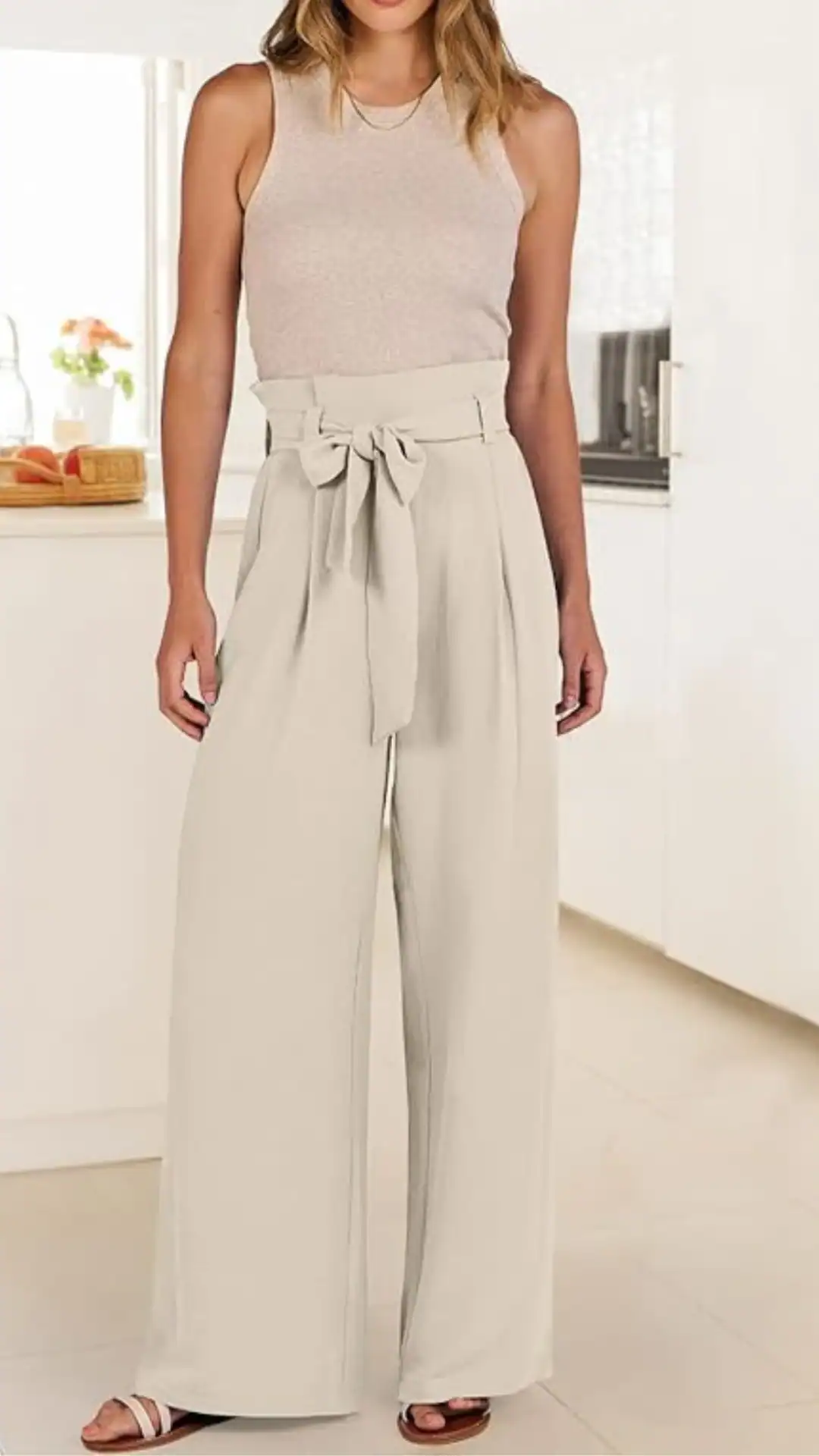 A model showcases high-waisted, wide-leg trousers in a soft beige fabric, featuring a belted waist and subtle pleating for a tailored appearance. Paired with a fitted, light taupe sleeveless top, this look exudes a minimalist and modern vibe, suitable for casual outings or brunch events.