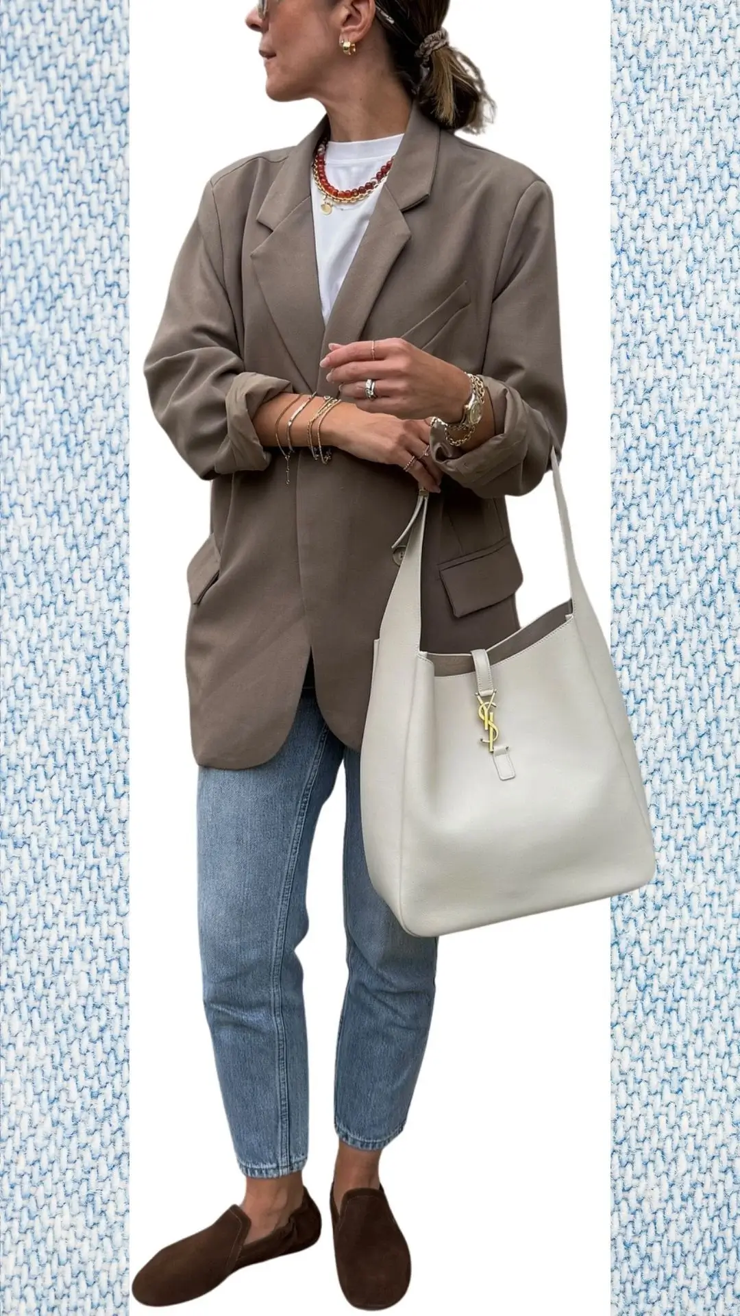 The outfit features light blue jeans made from a soft denim fabric with a slightly distressed finish. Paired with an oversized beige blazer in a warm, textured wool blend, the look is completed with a classic white t-shirt and brown suede slip-on shoes, accented by layered gold and red necklaces and a chic cream handbag with a distinctive gold charm. This stylish ensemble is perfect for layering during the colder months while transitioning into spring.