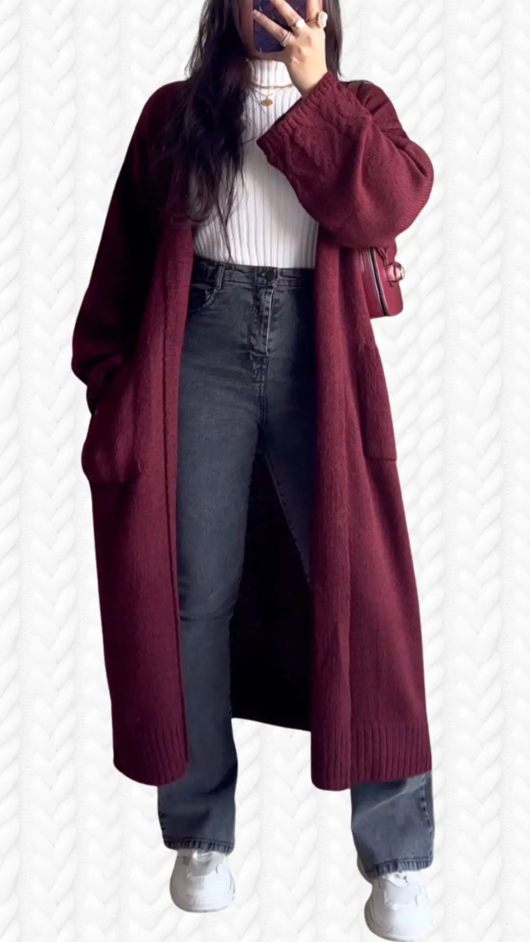 A long, burgundy cardigan made from a soft, textured fabric drapes elegantly over a white ribbed turtleneck, styled with high-waisted black jeans. The cardigan features wide, cozy sleeves and deep pockets, complemented by casual white sneakers for a chic, comfortable look. The outfit combines rich color tones and varied textures, creating a warm and inviting ensemble.