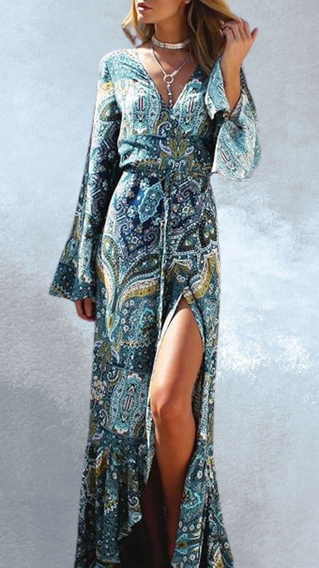 A fitted, long maxi dress features a vibrant paisley pattern in shades of teal, blue, and gold, capturing a bohemian country western vibe. The dress has flowing bell sleeves, a plunging neckline, and a thigh-high slit, which elegantly pairs with cowboy boots for a stylish country-chic look.