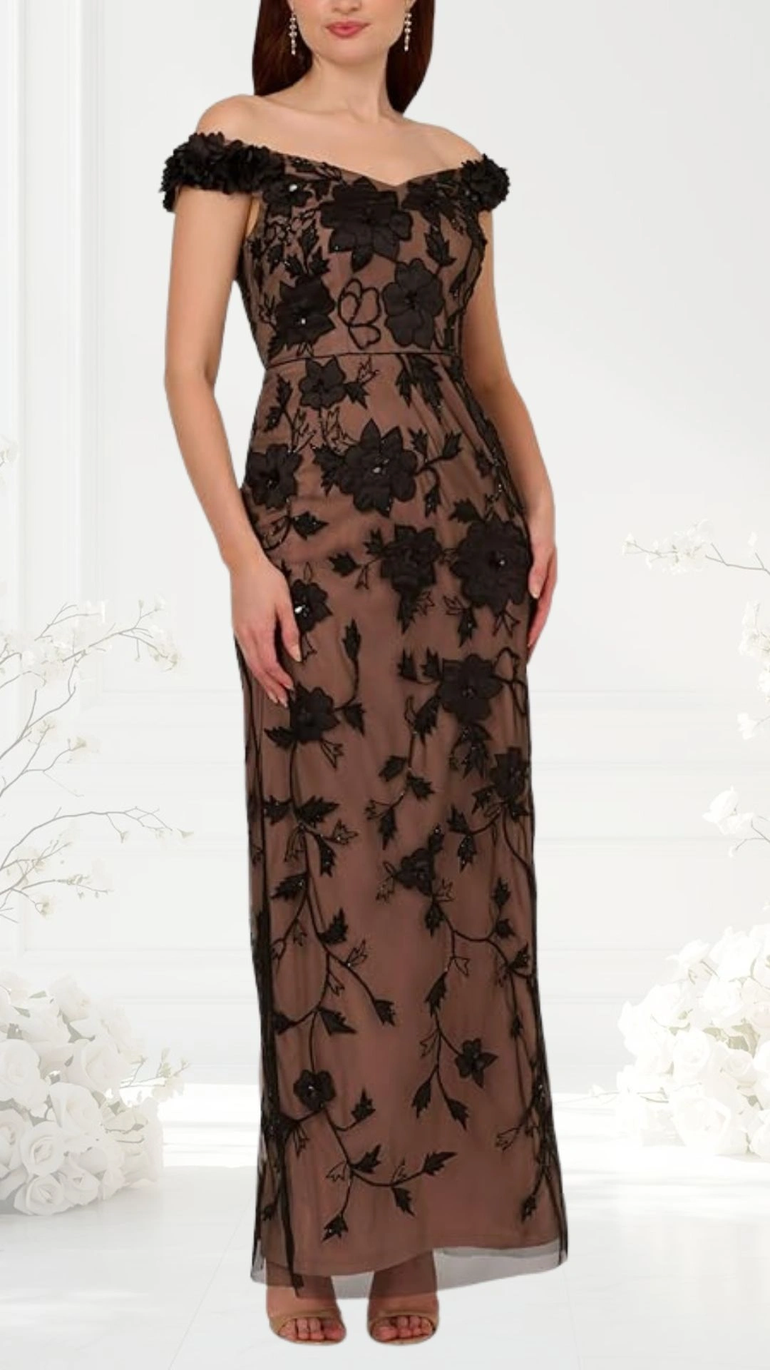 An elegant off-the-shoulder wedding guest dress features intricate black floral embroidery on a sheer, nude fabric. The fitted bodice transitions into a flowing skirt, offering a flattering silhouette. Styled with delicate embellishments, the dress is complemented by subtle, strappy heels.